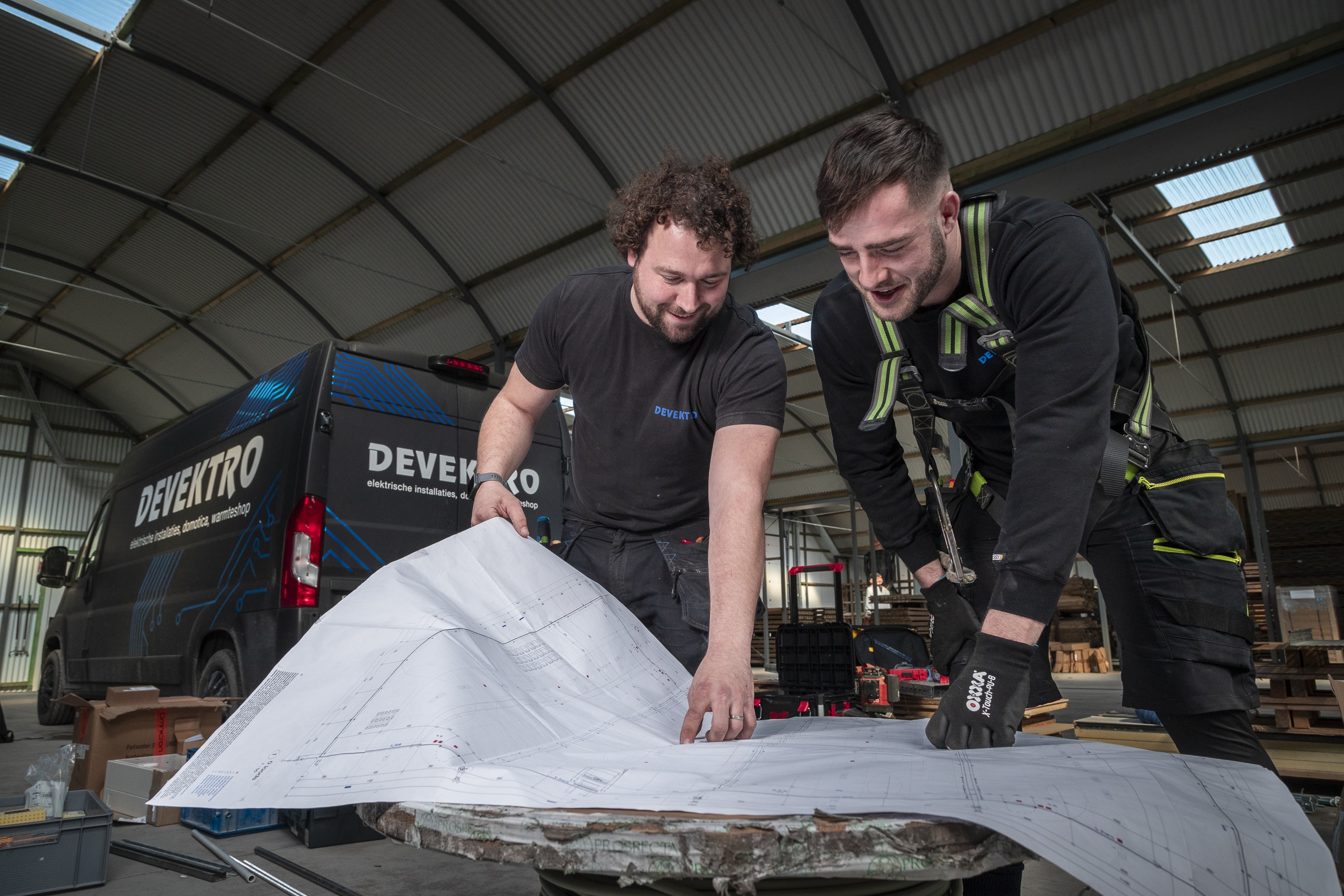 Twee mannen die voor een camionette staan en een technische tekening aan het bespreken zijn, klaar om te werken.